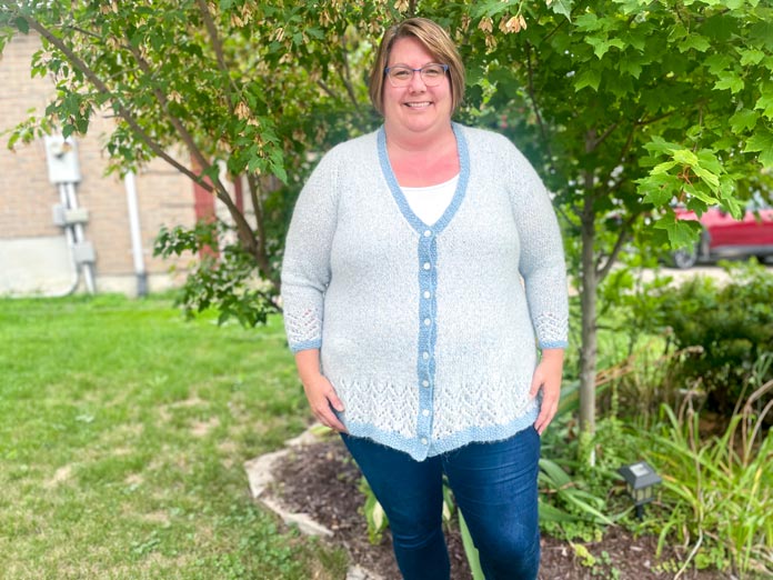 A woman wearing Lace Tipped Cardigan knitted with UNIVERSAL YARN Thula in Delphinium with blue trim knitted with UNIVERSAL YARN Fawning in Moonstone and UNIVERSAL YARN Penna in Clouded stranded together and Elan 15 mm white iridescent buttons
