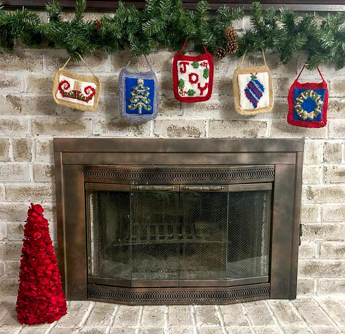 Five knitted Christmas ornaments hanging from a garland in front of a brick fireplace.