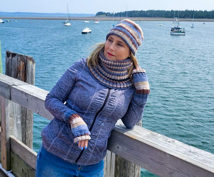 Michelle Porter is leaning on the rail of a wharf, with the ocean and boats in the background, wearing a blue and brown striped hat, mitts, and cowl, knit from UNIVERSAL YARN Deluxe Stripes, color Timber.