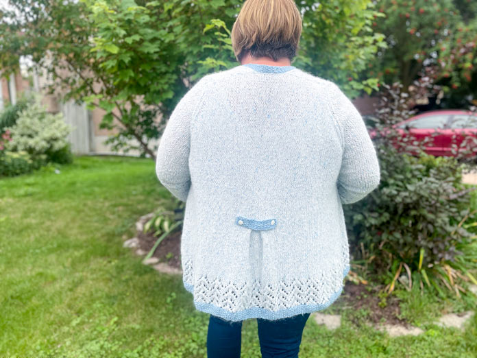 A woman wearing Lace Tipped Cardigan knitted with UNIVERSAL YARN Thula in Delphinium with blue trim knitted with UNIVERSAL YARN Fawning in Moonstone and UNIVERSAL YARN Penna in Clouded stranded together and Elan 15 mm white iridescent buttons.