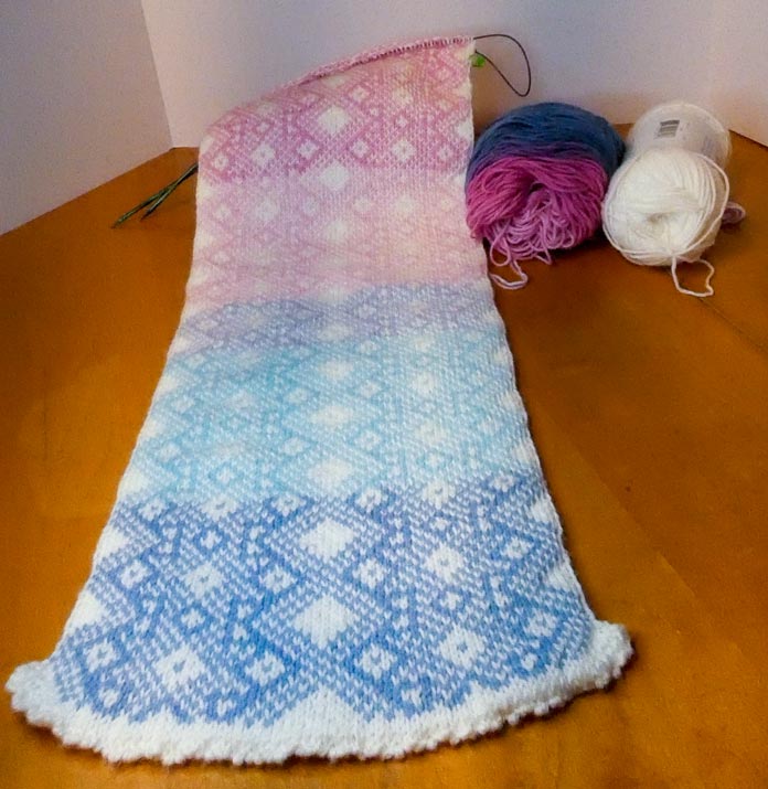 A section of the scarf, showing a colored pattern on a white base. The pattern is a Nordic star design that repeats twice across each row. The striping of the yarn creates subtle color changes from blue to pale blue, mauve, then pale and light pink. The remaining yarn sits beside the work, showing the end of the cake with medium pink and denim blue remaining, with the ball of solid white to the right of that.