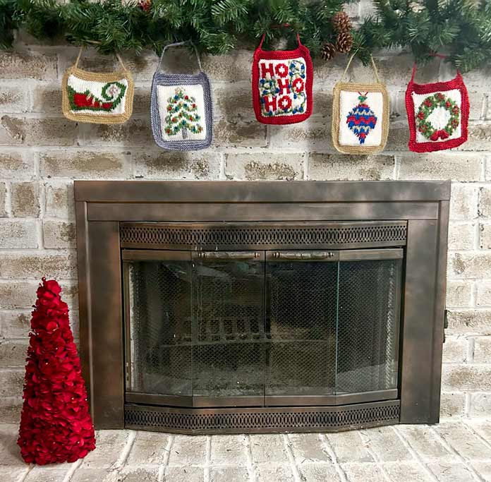 Five knitted Christmas ornaments hanging from a garland in front of a brick fireplace.