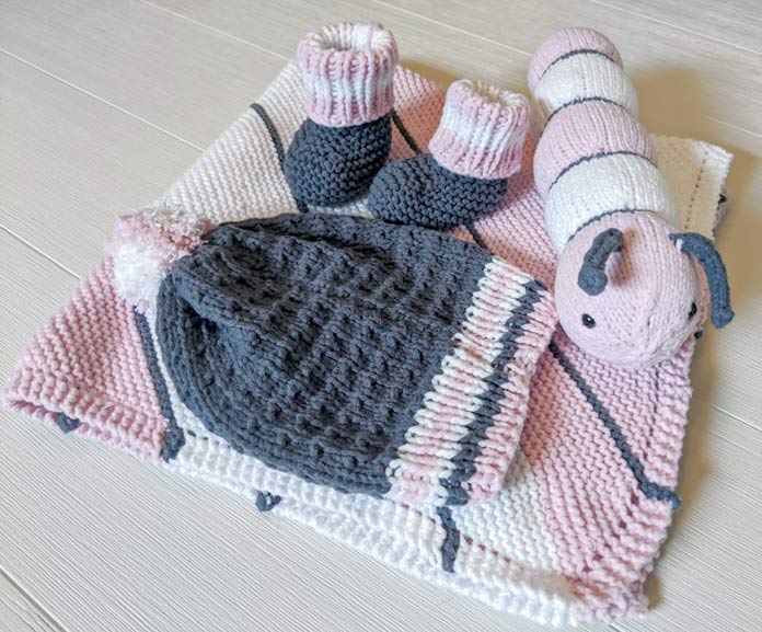 The I Want to Knit a Baby Blanket, Pompom Baby Hat, Booties, and Cuddly Caterpillar made with Feels Like Butta in pink, white, and gray stripes are displayed together on a white wooden table.